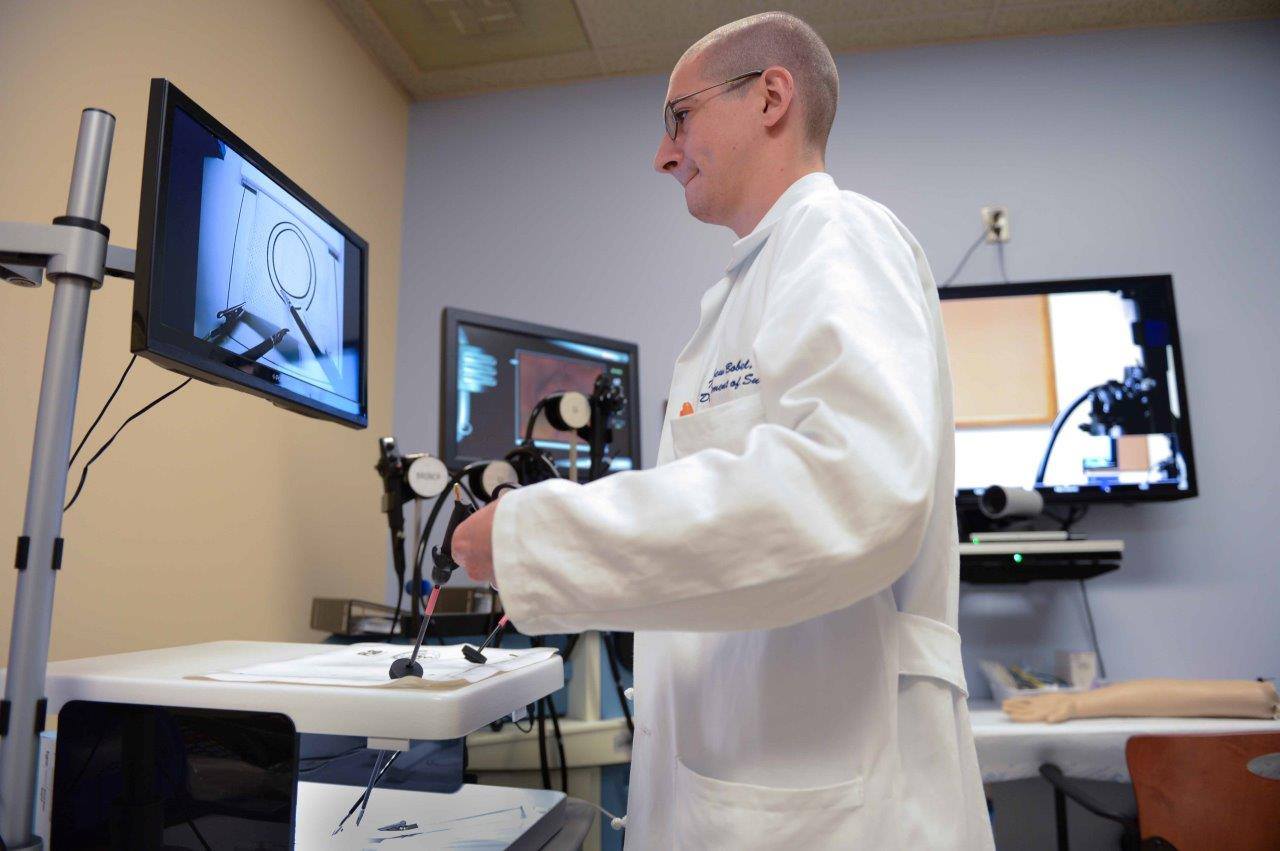 Image of a person with glasses, short hair and a white lab coat using simulation equipment and a computer monitor