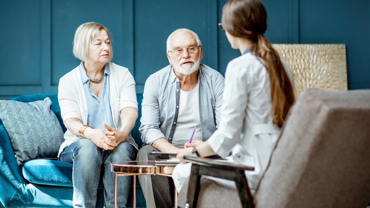 Two seniors sitting on couch talking to a nurse.