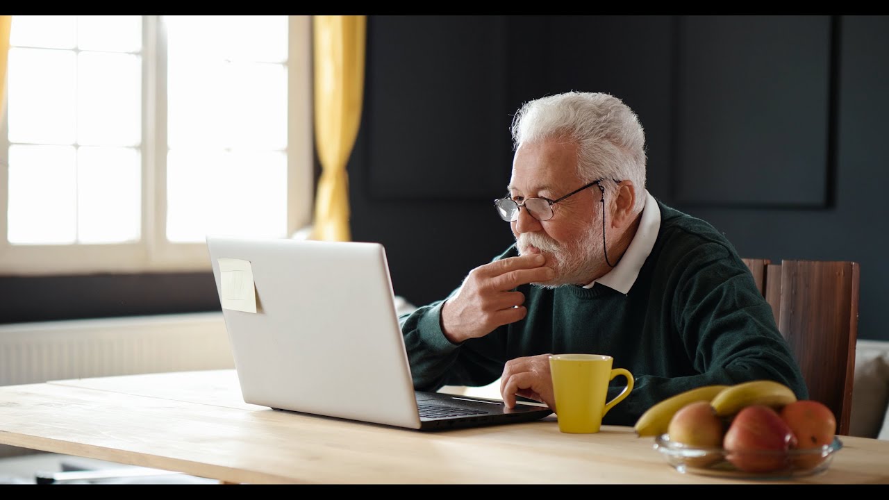 Senior man at a desk using a laptop with fruit and coffee.