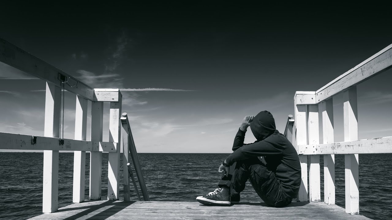 Black and white photo of a person in a hoodie sitting on a pier.