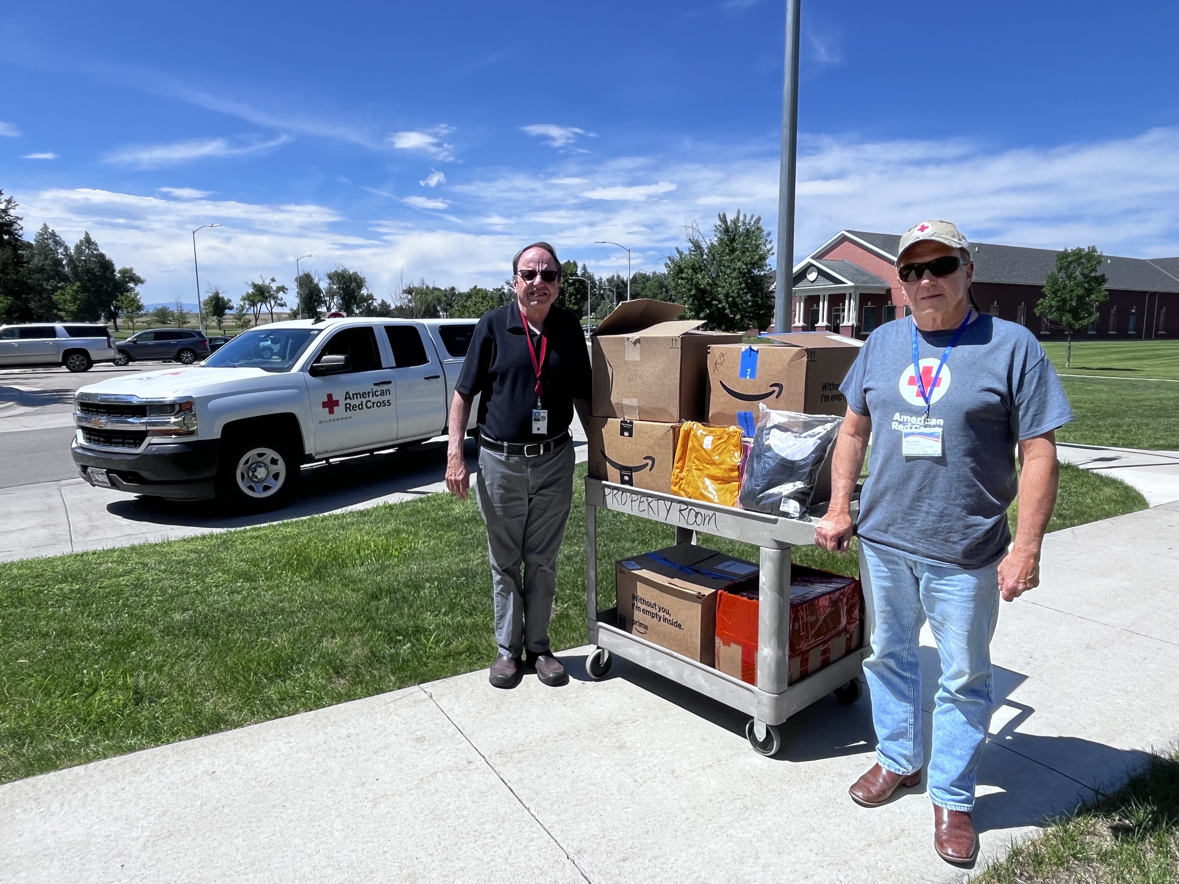 Two men stand by a donation cart with boxes, in front of a white pickup truck.
