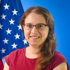 A woman in a maroon shirt seated in the foreground with an American flag and blue background.