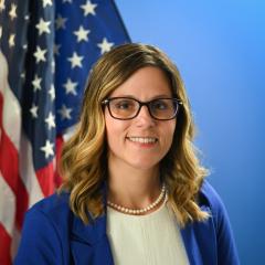 A woman in a blue jacket and white shirt with an American flag in the background.