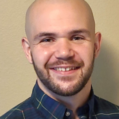 man with beard, wearing a dark blue button up shirt.  background is a beige wall.