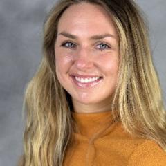 Woman with long blonde hair, wearing an orange blouse.  Background is grayish cloud-like backdrop.