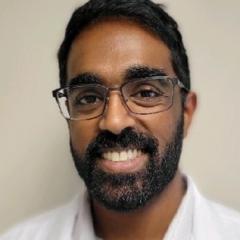 man with black hair, beard and mustache, wearing glasses and white button down polo-style shirt.  Background is a cream colored wall.