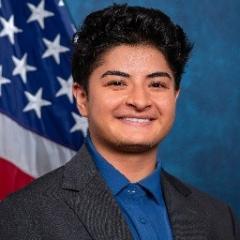 Young adult with black hair wearing blue shirt and a dark blue or black blazer.  Background standard blue with American flag.