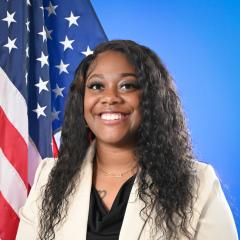 A smiling woman posed in front of an American Flag and blue background.