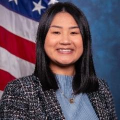 Young woman with black hair, light blue blouse, black and white tweed blazer, standard blue background with American Flag.