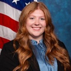 Young woman with long red hair, light blue blouse, dark blue / black blazer.  Background of standard blue with American flag.