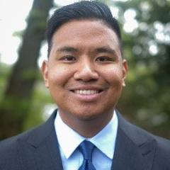 Young man with black hair in light blue shirt, blue tie, dark blue blazer with white pocket square.  Background is outdoors with trees and foliage