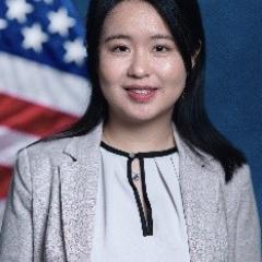 Young woman with black hair, white blouse, light gray blazer.  Background standard blue with American Flag.