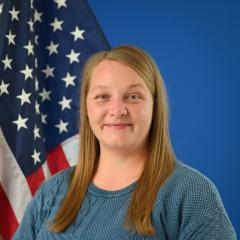 A smiling blonde woman posed in front of an American flag and blue background.