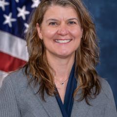Woman with shoulder-length wavy brown hair smiling in front of an American flag.