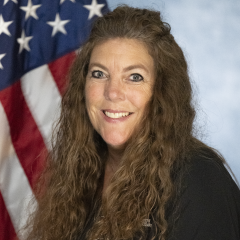Woman with long brown hair, wearing black shirt, smiling in front of American flag.