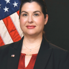 Woman in black blazer and red shirt with American flag in background.