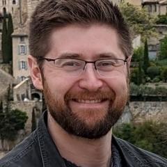 Photo of man with brown hair, brown beard, and glasses smiling in front of a town.