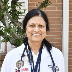 Smiling woman in white lab coat with stethoscope, standing by a tree.