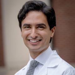 Smiling man in white lab coat and gray tie stands against brick wall.