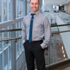 Man in gray shirt and blue tie standing in office building with hands in pockets.