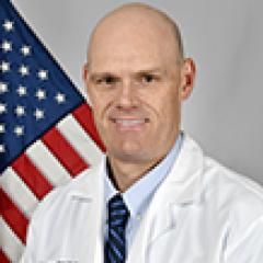 A bald man in a white lab coat and blue tie stands in front of an American flag.