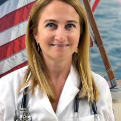 Woman with blonde hair in white lab coat with stethoscope around her neck in front of an American flag.