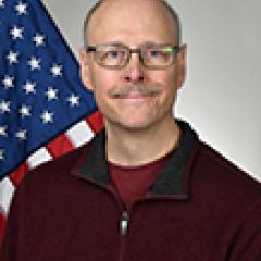 Man with mustache wearing glasses and maroon sweater in front of US flag.