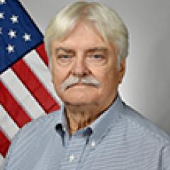 Older man with white hair and mustache, wearing blue shirt, stands in front of American flag.