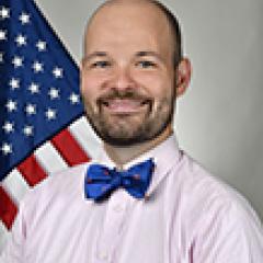 Man with a pink shirt and blue bowtie stands in front of the American flag.