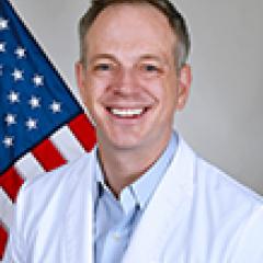 A smiling doctor in white coat with American flag in the background.