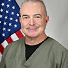 Man in green scrubs and black shirt underneath, smiling with American flag behind him.