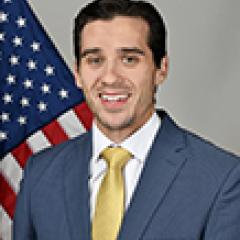 Man in blue suit and yellow tie smiles in front of American flag.