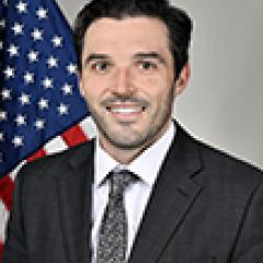 Man in a suit and tie smiling with an American flag behind him.