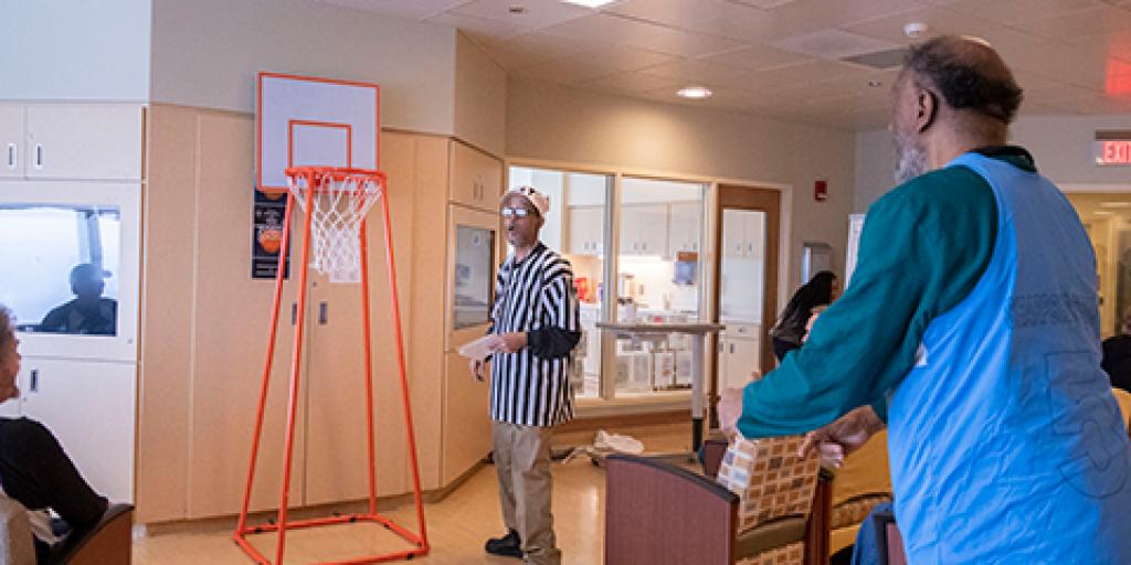 A Veteran shoots a small paper basketball towards a hoop while a referee watches