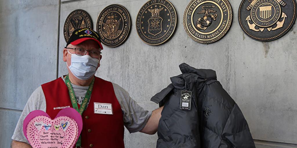 Dan Condron wears his Red Coat Ambassador vest and 1st Marine Division hat as he stands in the lobby of building 3 at Coatesville VA Medical Center and holding out a valentine and a coat while welcoming Veterans on February 16, 2022