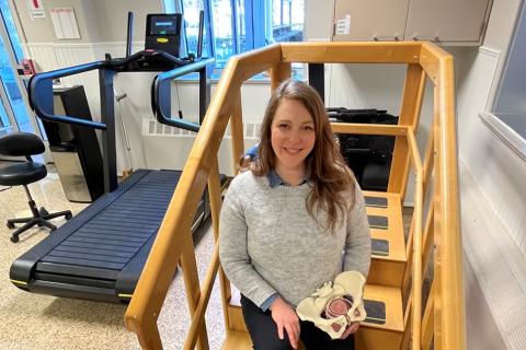 Female seated on therapeutic steps near a treadmill holding a 3D skeletal model of a pelvis